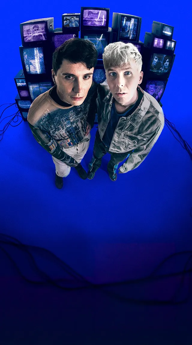 Dan and Phil are stood in front of a stack of old analogue TVs displaying videos from their channel. The image is shot with a fisheye lens and they are styled in late-80s clothing.